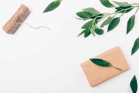 Top view decorative frame of greenery, twine and box wrapped in brown eco paper, copy space. Flat lay arrangement of natural green leaves and gift on white background with empty place.の写真素材
