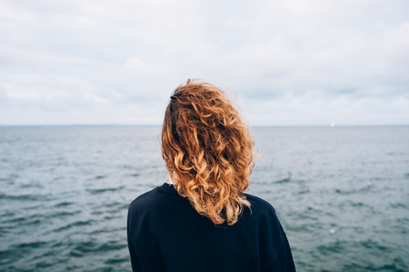 Female with red curly hair outdoors standing alone thinking near blue water. Rear view of young woman looking at sea.の写真素材