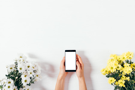 Flat lay festive composition woman's hands holding mobile phone between two bouquets of flowers on white wooden table, top view with copy space.の写真素材