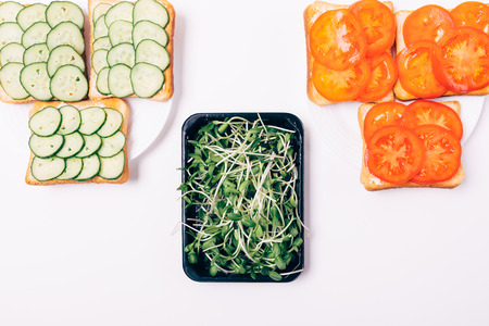 Two types of sandwiches with cucumbers and tomatoes next to box of sprouted sunflower seeds, top view.の写真素材