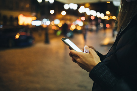 Close-up woman's hands using mobile phone with glowing screen on street, lights of night city on background.の写真素材