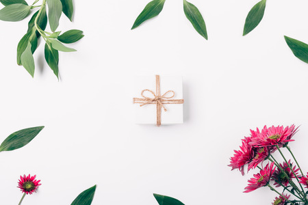Small gift box in center of flat lay floral frame made of fresh green leaves and blooming pink flowers on white background, top view.の写真素材