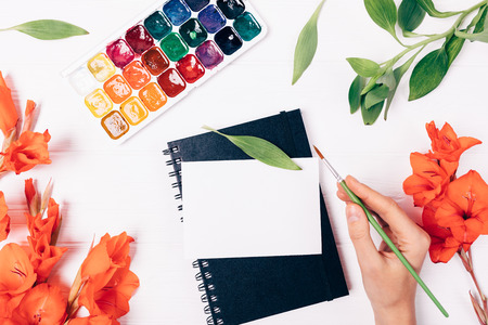Woman's hand holding brush near postcard, sketchbook and watercolor paints surrounded by lush red flowers and leaves on white background, top view.の写真素材