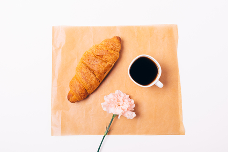 Flat lay feminine composition of simple breakfast of croissant and cup of black coffee with pink flower on white background, top view.の写真素材
