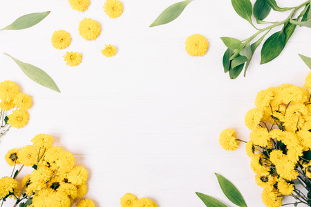 Festive floral flat lay frame of yellow flowers and green leaves on white wooden table with blank center, top view.の写真素材