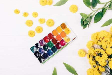 Decorative flat lay composition palette of watercolor paints, bright yellow flowers and green leaves on white wooden table, top view.の写真素材