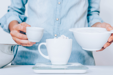 Close-up flour in white cup. Young woman wearing blue shirt weighing baking ingredient using scales.の写真素材
