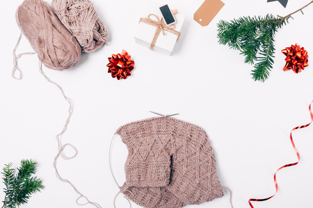 Flat lay composition handmade knitted cloth made of natural wool next to gift box, Christmas red ornaments and fir branch, top view on white table.の写真素材