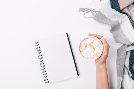 Flat lay composition female's hand holding mug of coffee with marshmallows near open blank notepad and checkered plaid on white background, view from above.の写真素材