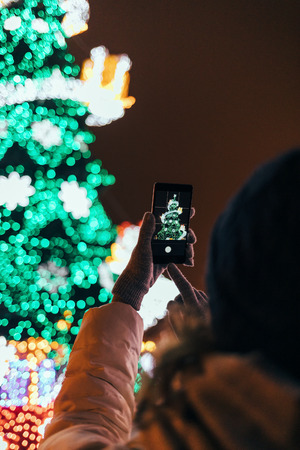 Woman's hands in warm gloves using mobile phone photographing illuminated green Christmas tree in...