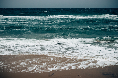 Beautiful seascape in rough weather, foamy waves roll on sandy beach. Gloomy day with heavy dark sky.の写真素材