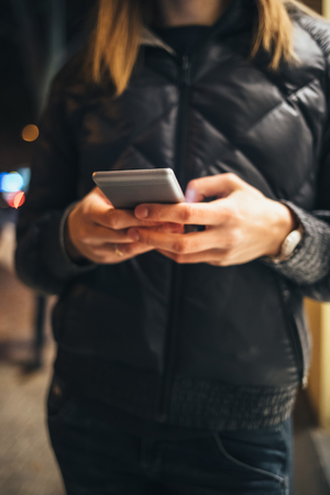 Young woman typing on mobile phone on street in city at winter night, close-up.の写真素材
