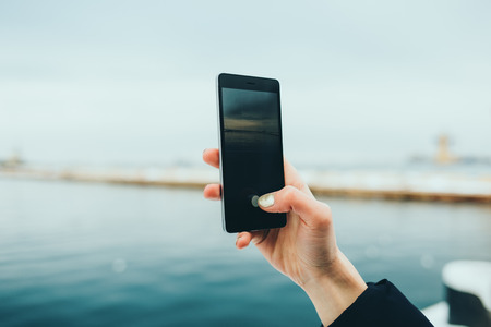 Close-up of female hand holding mobile phone photographing seascape on winter day.の写真素材