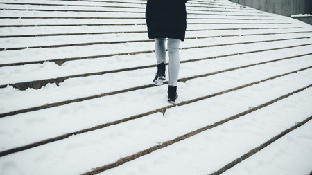 Young woman wearing winter coat and boots walking up on stairs covered with snow. Female's legs stepping on slippery steps outdoors.の写真素材