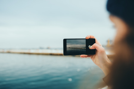 Candid image of female hand holding smart phone photographing sea on cloudy winter day, close-up.の写真素材