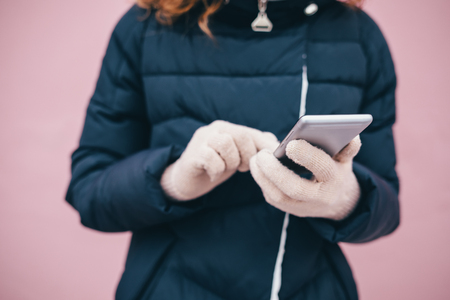 Close-up of young woman's hands in warm gloves using mobile phone outdoors in winter on pink wall background.の写真素材