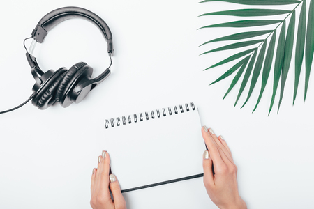 Woman's hands holding blank notepad near black headphones and green palm leaf on white table, top view.の写真素材
