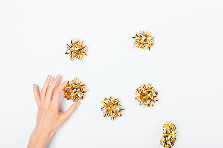 Female's hand holding one of golden bows for wrapping gifts on white background, top view. Festive flat lay arrangement concept of preparation for holidays.の写真素材