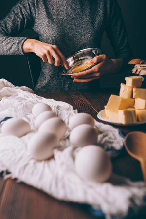 Process of cooking dough for pie. Cropped image of woman mixing sugar and yolks in metal bowl next to eggs, diced butter and jar of jam on brown wooden table.の写真素材