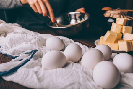 Close-up female's hands whisking ingredients in metal bowl next to diced butter and eggs on brown wooden table. Process of cooking dough for homemade cake.の写真素材