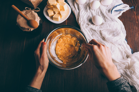 Process of cooking dough for pie. View from above female's hands mixing sour cream, sugar and yolks in metal bowl next to eggs, diced butter and jar of jam on brown wooden table.の写真素材