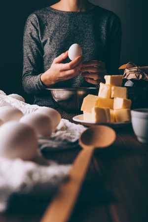 Cropped photo of young woman holding egg next to ingredients for baking homemade pie: diced butter and jam. Process cooking dough at home.の写真素材