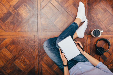 Young woman working from home writing in notepad near headphones and mug of coffee. Female's legs in jeans on wooden floor, copy space. Concept comfortable freelancing.の写真素材