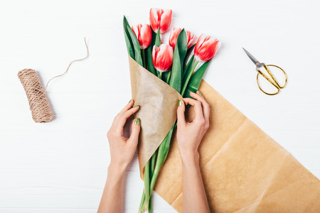 Woman's hands wrapping bunch of red tulips in brown eco paper next to scissors and jute rope on white wooden table, top view. Flat lay composition of making bouquet of fresh flowers.の写真素材