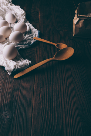 View from above ingredients for cooking cake on brown wooden table: eggs on white towel and paper bag of flour next to spoons.の写真素材