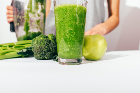 Close-up glass of fresh made healthy green smoothie at white table near ingredients: apple, broccoli and celery. Young woman making wholesome drink in blender.の写真素材