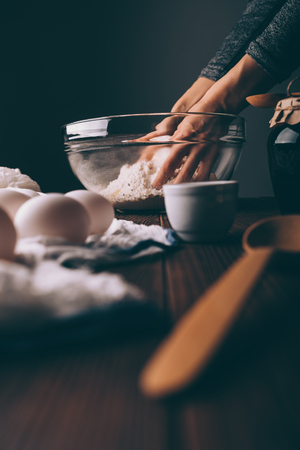 Close-up of female's hands kneading dough in big glass bowl on brown wooden kitchen table near eggs...