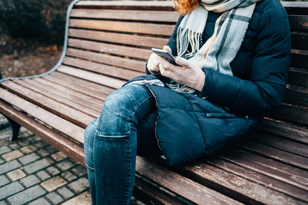 Close-up young unrecognizable woman wearing warm coat and jeans sitting on bench in city using smart phone on cold winter day.の写真素材