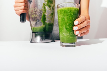 Woman's hands with white manicure holding blender and glass of freshly made green smoothie on kitchen table, close-up.の写真素材