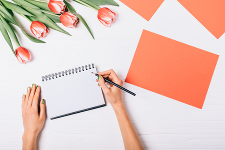 Woman's hands writing in empty notebook next to blank red sheet of paper and bouquet of beautiful fresh tulips, top view. Flat lay floral arrangement concept of spring greetings.の写真素材