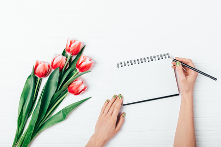 View from above of woman's hands writing in blank open notepad and bouquet of fresh tulip flowers on white wooden desk, flat lay.の写真素材