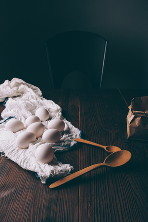 Ingredients for cooking cake on brown wooden table: eggs on white towel and paper bag of flour next to spoons.の写真素材