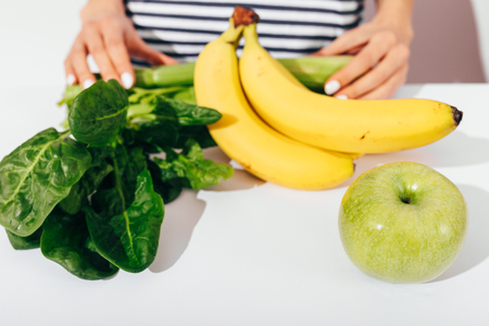 Close-up young woman in striped t-shirt standing at white table with fresh and healthy fruits and vegetables: bananas, apple, spinach leaves and celery.の写真素材