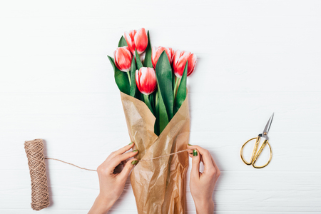 View from above woman's hands wrapping bouquet of freshly cut red tulips into brown kraft paper on white wooden table, flat lay.の写真素材