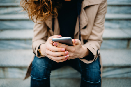Close-up young woman wearing beige trench coat and blue jeans sitting on gray stone stairs using smart phone. Female's hands holding mobile device and typing text messages outdoors in city.の写真素材