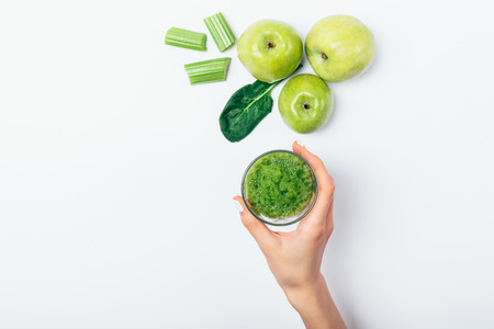 Top view female's hand holding glass of green smoothie next to ingredients: fresh apples, spinach and celery on white background, flat lay.の写真素材