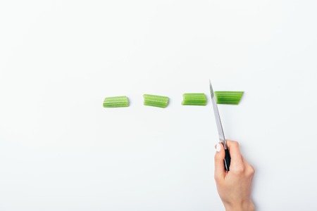 Female's hand cut the celery stick with knife on white table, top view. Minimal flat lay composition...