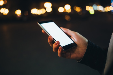 Close-up woman's hand holding smart phone with blank white screen in city at night on background of blurred street lights.の写真素材