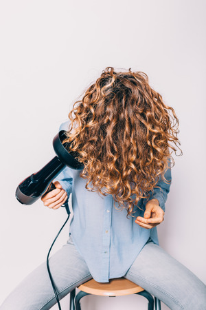 Young woman sitting on chair styling her curly hair with hairdryer with special diffuser nozzle.の写真素材