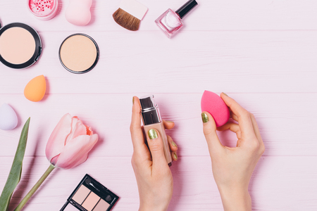 Woman's hands holding nude beige foundation and makeup sponge next to decorative cosmetics on wooden table, top view. Flat lay composition of beauty products and accessories.の写真素材
