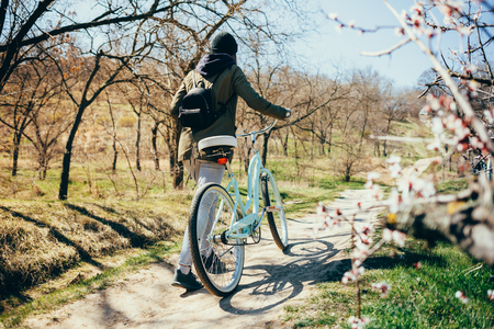 Young hipster woman wearing jacket, hat and backpack walking next to her bike along footpath in park on sunny spring day.の写真素材
