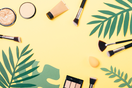 Women's makeup products for smooth skin tone and bronze tan next to palm branches on yellow background with empty space in center, top view.の写真素材