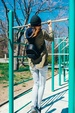 Rear view of young woman in casual clothes with backpack doing pull-ups on horizontal bar on sports...