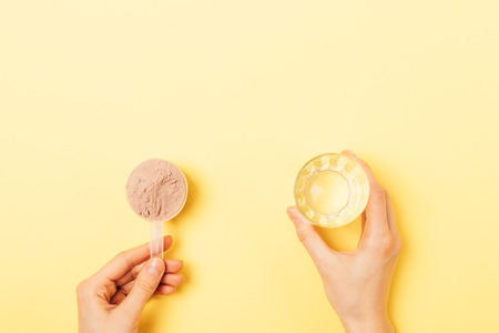 View from above woman's hands holding chocolate protein powder in scoop and empty glass on yellow background with copy space.の写真素材