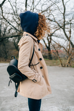 Stylish fashionable young woman dressed in beige trench coat and hood sweatshirt with backpack standing in city park holding hands in pockets, side view.の写真素材