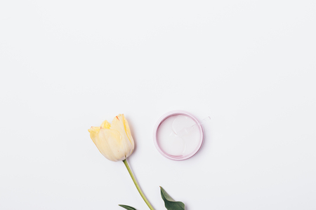 Jar of cosmetic patches for skin under eyes next to delicate tulip flower on white background, minimal flat lay composition.の写真素材